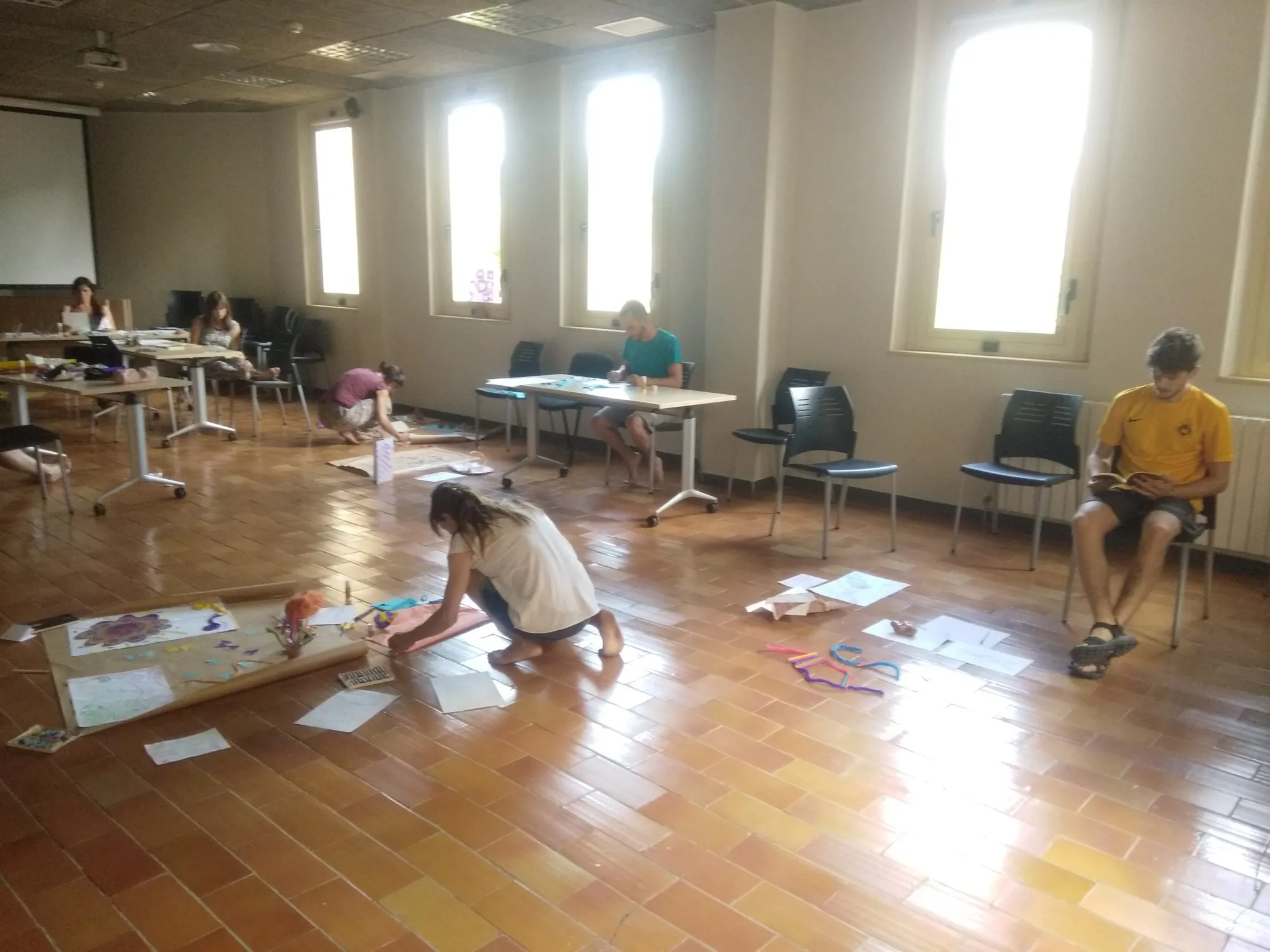 Mestres i educadores treballant a terra i en taules en una sala amb finestres altes.