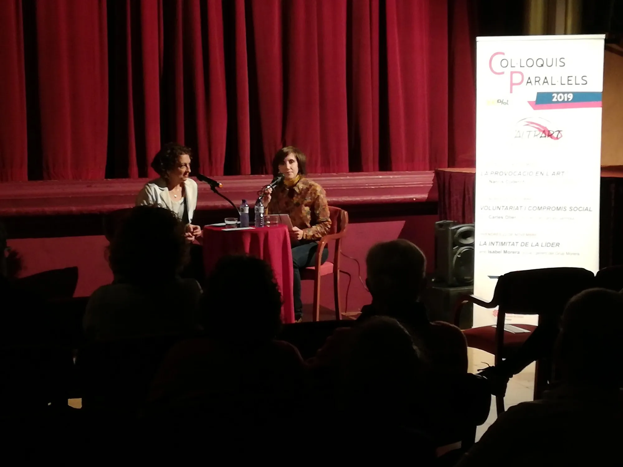 Entrevista a Isabel Morera, amb el cartell dels Col·loquis Paral·lels al costat.