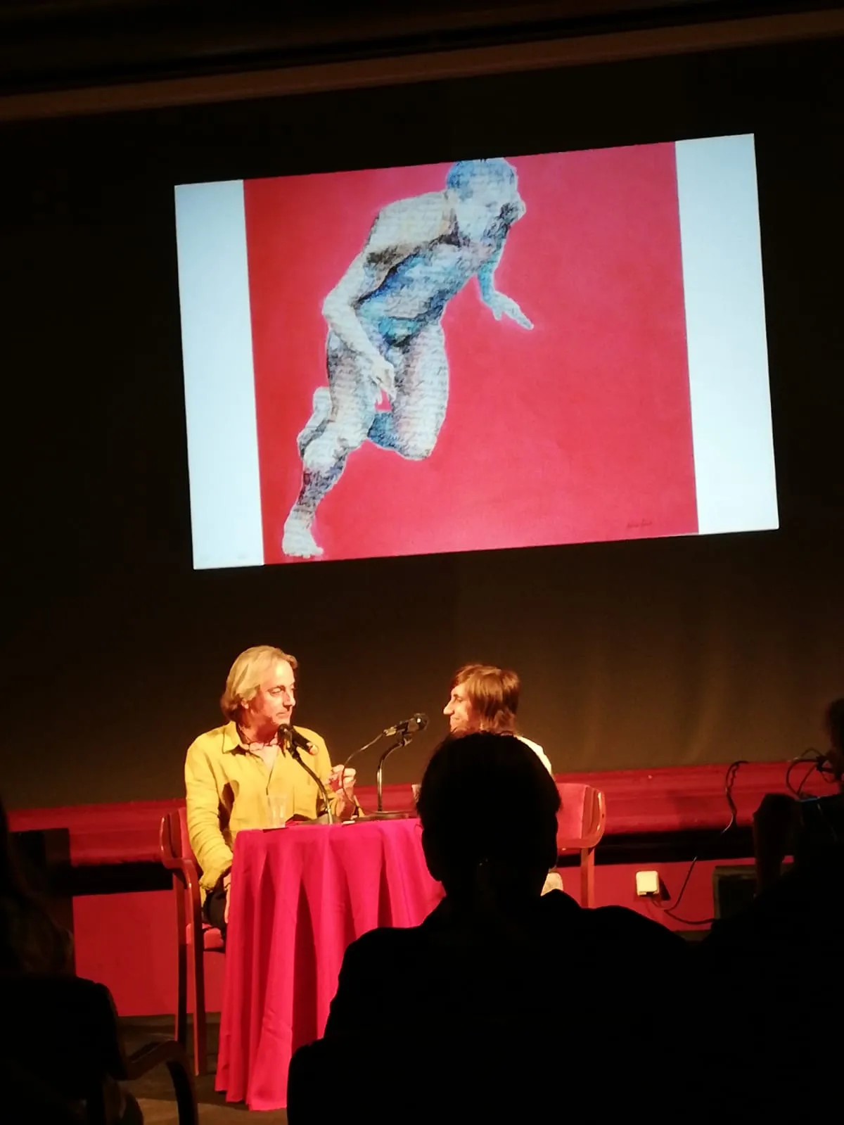 Narcís Coderch parla davant d'una projecció d'una figura humana en moviment sobre fons vermell.