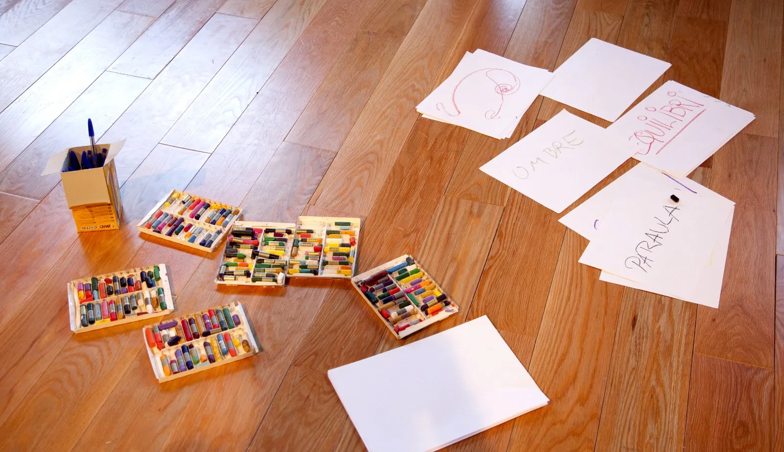 Capses de pastels i fulls amb dibuixos escampats sobre un terra de fusta.
