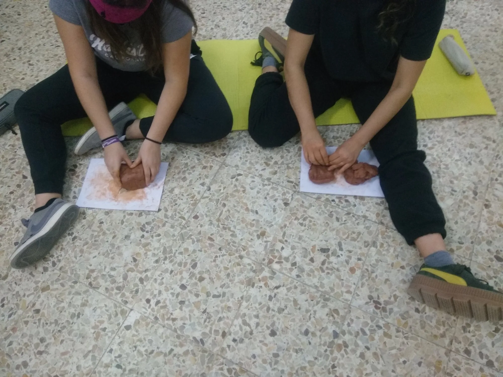 Dues alumnes assegudes a terra modelant fang sobre fulls de paper.
